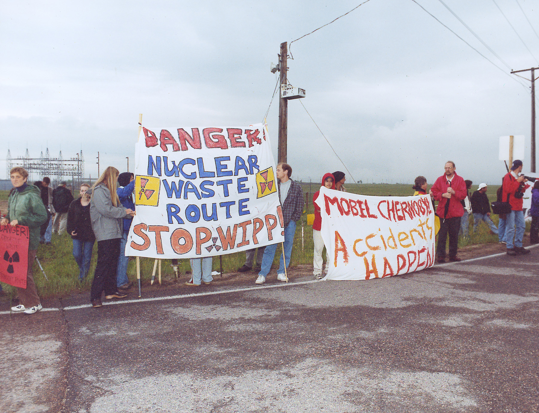 Group of mostly white-presenting activists stand beside a roadway with two handmade banners. One reads: Danger! Nuclear Waste Route. Stop WIPP. The other reads: Mobile Chernobyl, Accidents Happen.